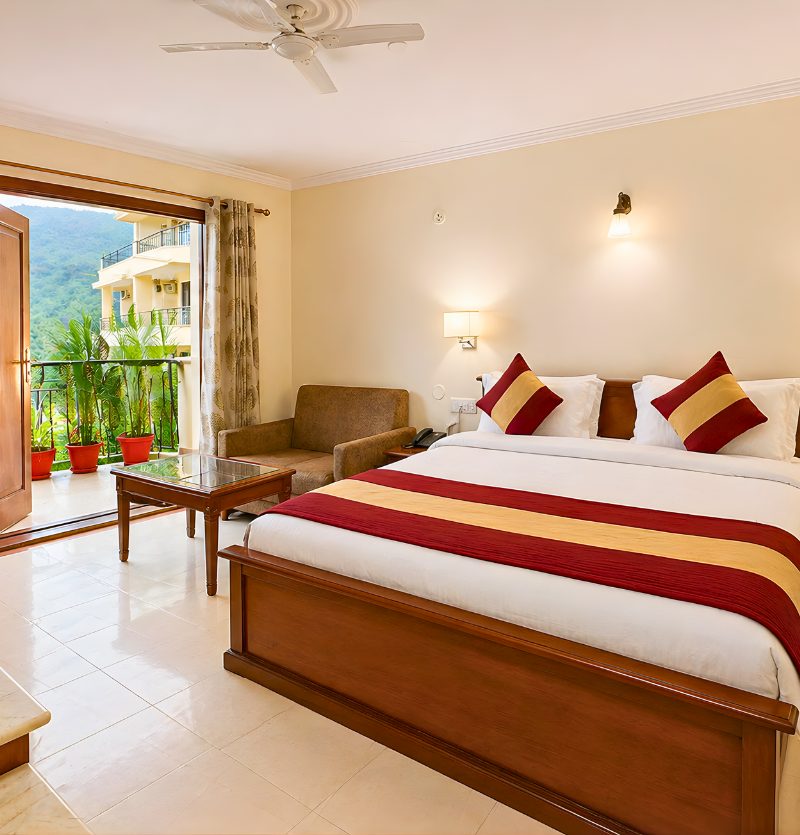 King-size bed with a red-gold throw and matching pillows in a bright hotel room, next to a balcony with potted plants.