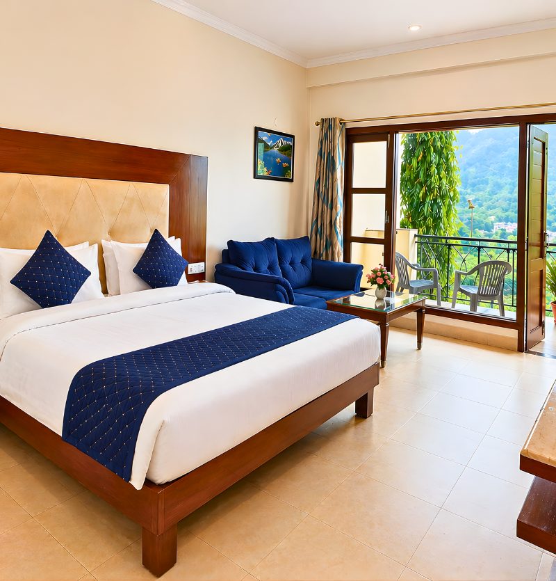 Hotel room with a large wooden bed, white linens, and blue accents; blue sofa, glass coffee table, and balcony with chairs overlooking a green mountainous view