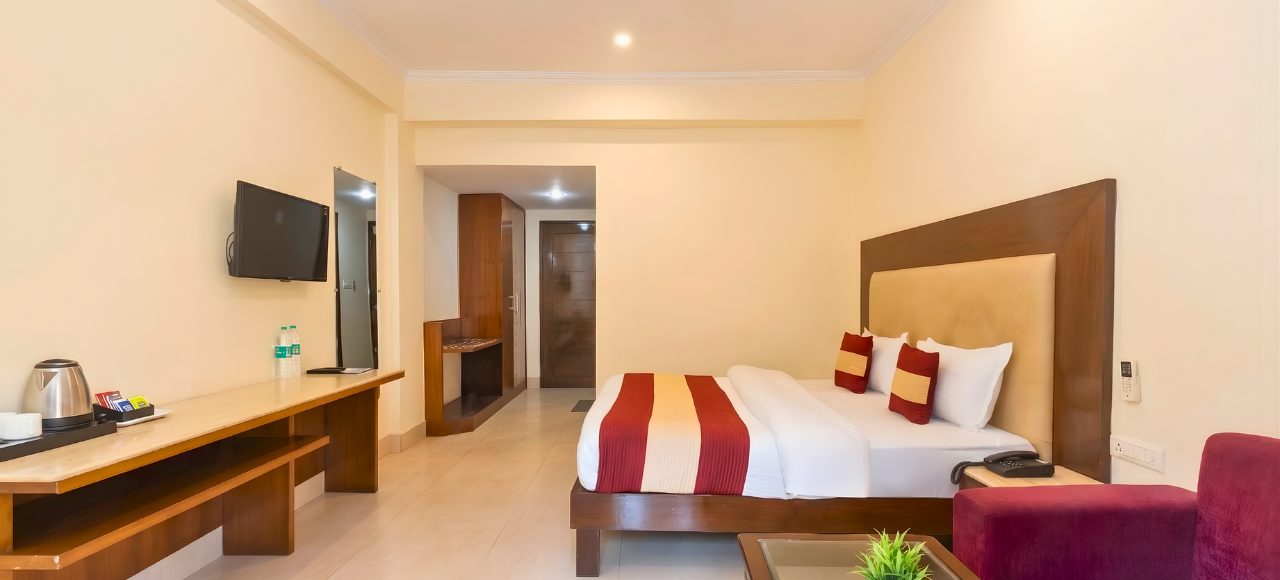 Hotel room with a large bed featuring a brown headboard, white linens, and red-gold striped runner; desk, wall TV, and purple sofa nearby.