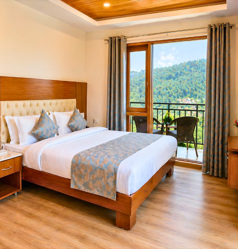 Hotel bedroom with a large wooden bed, tufted headboard, and blue patterned bed runner and pillows; glass doors open to a balcony with two chairs, overlooking green hills.