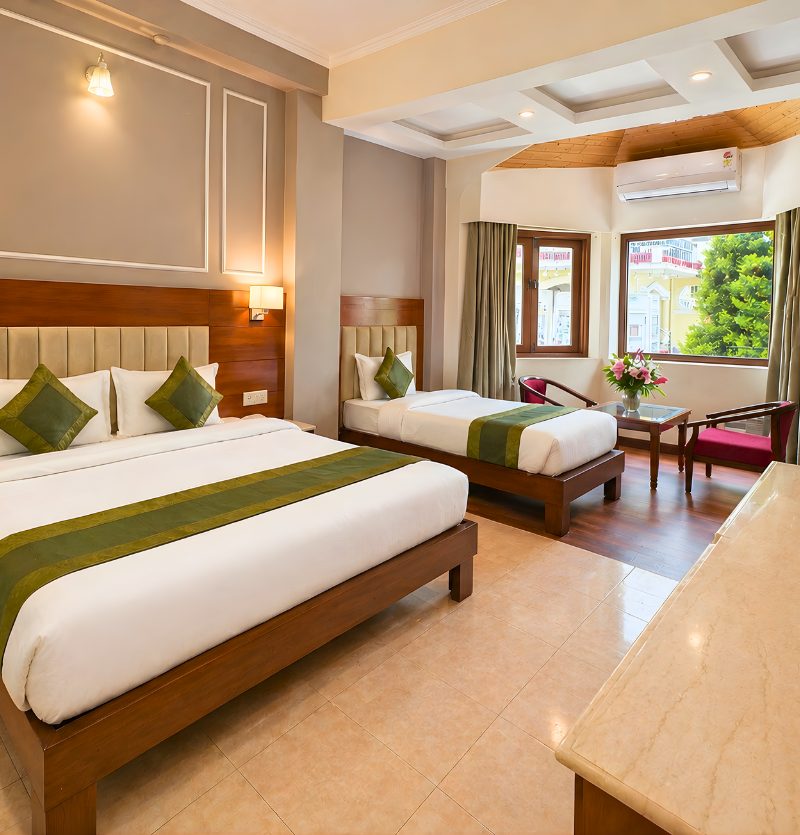 Hotel room with two wooden beds, white linens, and green accent pillows and runners, bright and modern décor. Window seating area and flowers nearby.