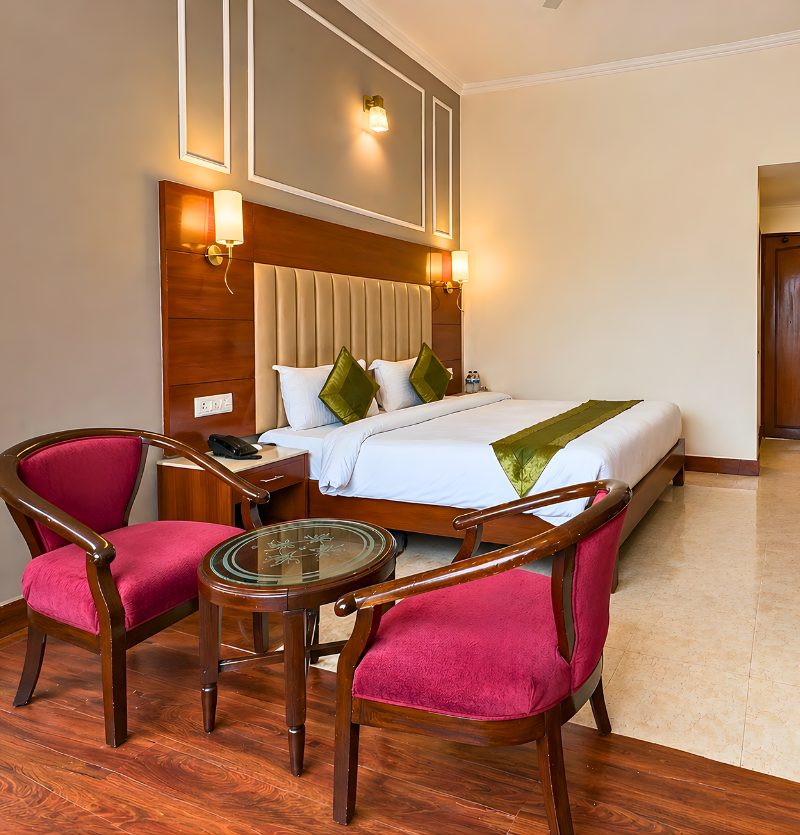 Hotel room with a large bed, two red chairs, and a glass-topped table in a wood-accented space.