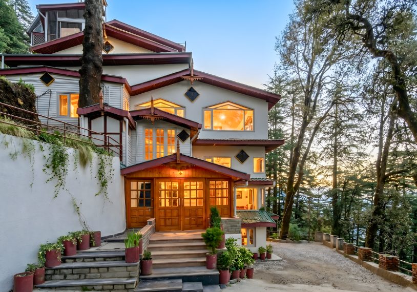Three-story hillside house with warm glowing windows, red roof accents, and potted plants along the entry stairs at sunset.