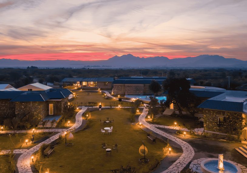 Evening view of a resort courtyard with winding stone paths, warm lights, and outdoor seating against a backdrop of distant mountains at sunset.