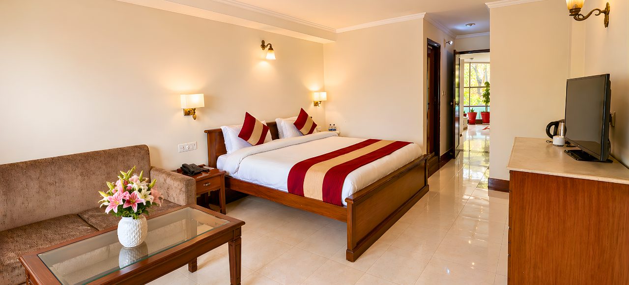 Hotel room with a large wooden bed, burgundy-beige runner, two pillows, brown sofa, glass coffee table, and a vase of pink lilies.