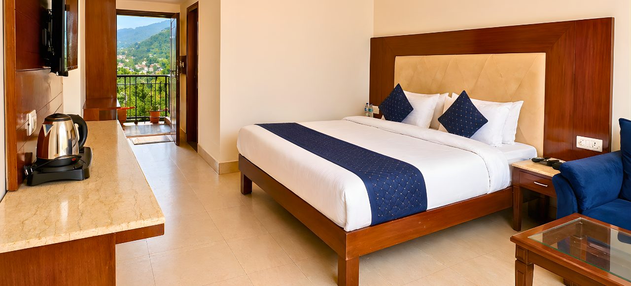 Hotel room with a king bed, white linens, navy accents, and an open balcony door showing mountains in the distance.
