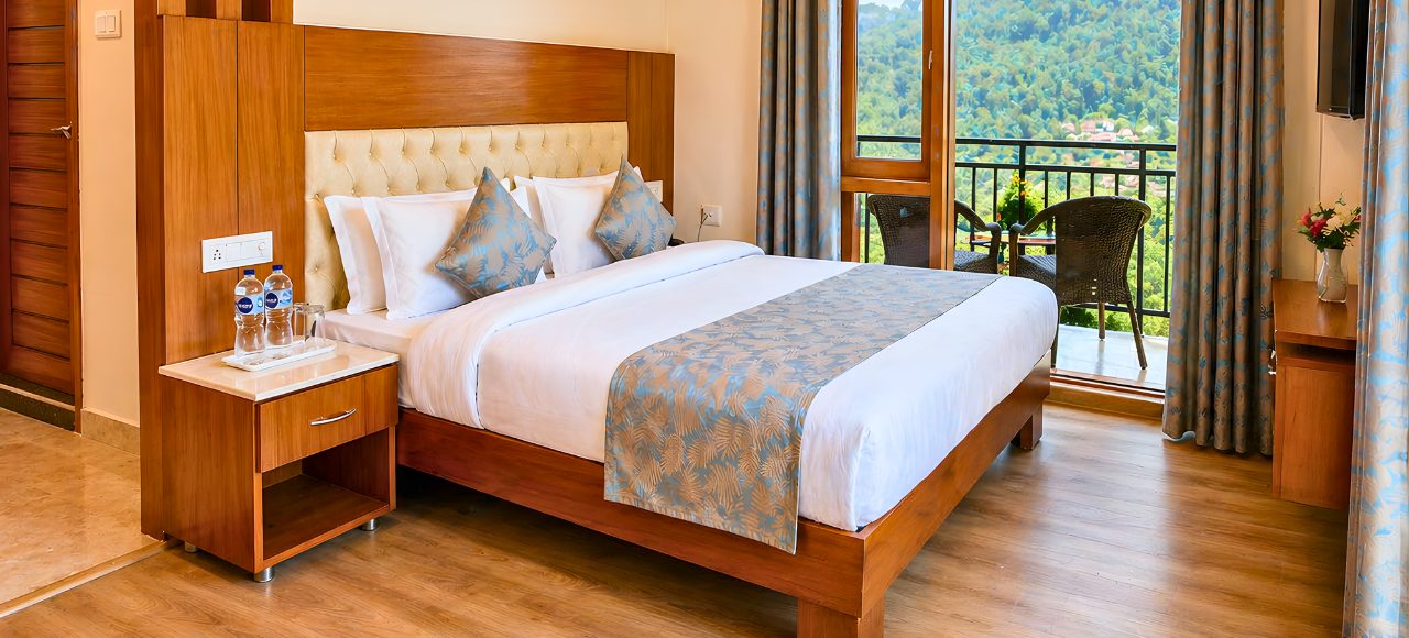 Hotel room with a large wooden bed, white linens, and blue patterned runners and cushions, next to a nightstand with water bottles and glasses.