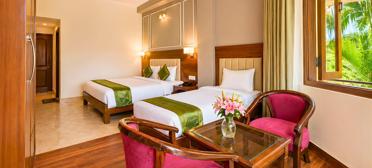 Hotel room with two twin beds, white linens, green runners, and a seating area by a window.