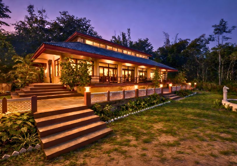 Modern single-story house with warm lights on, stairs and railing leading to a landscaped garden at dusk.