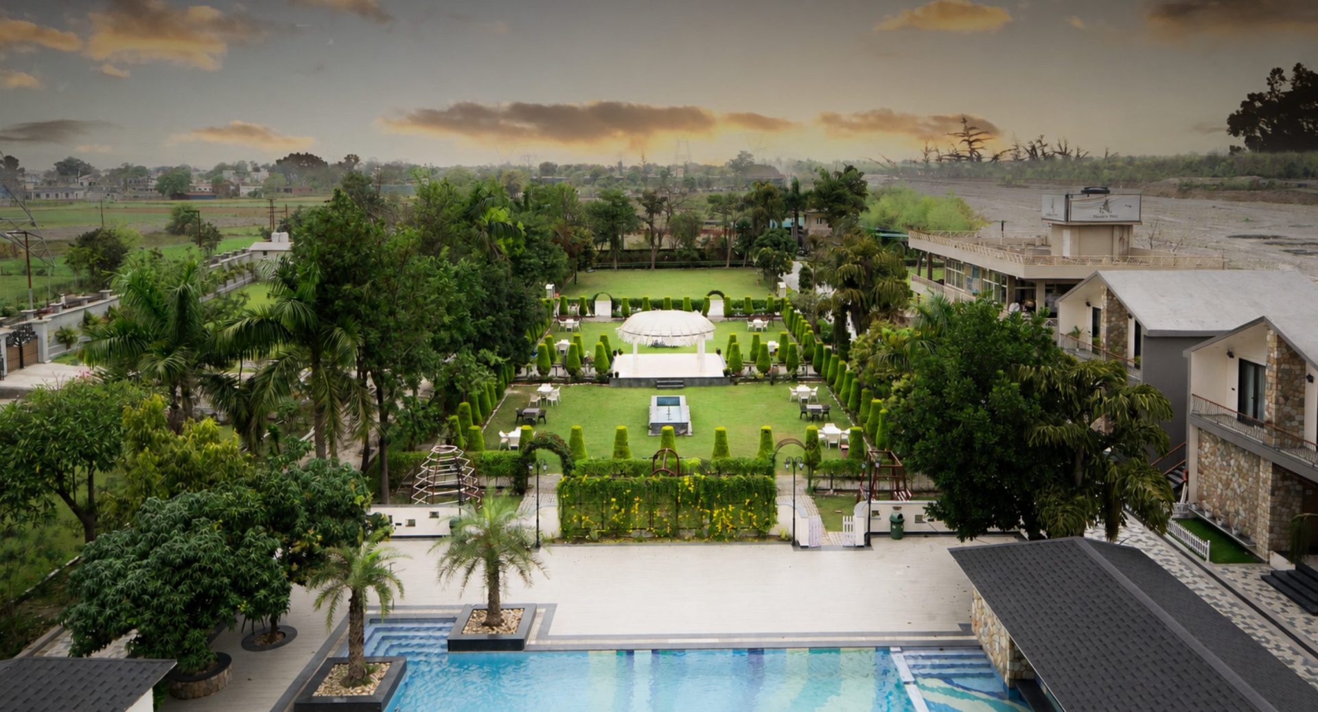 Aerial view of a resort with a rectangular pool, manicured gardens, and a central gazebo surrounded by chairs and paths.