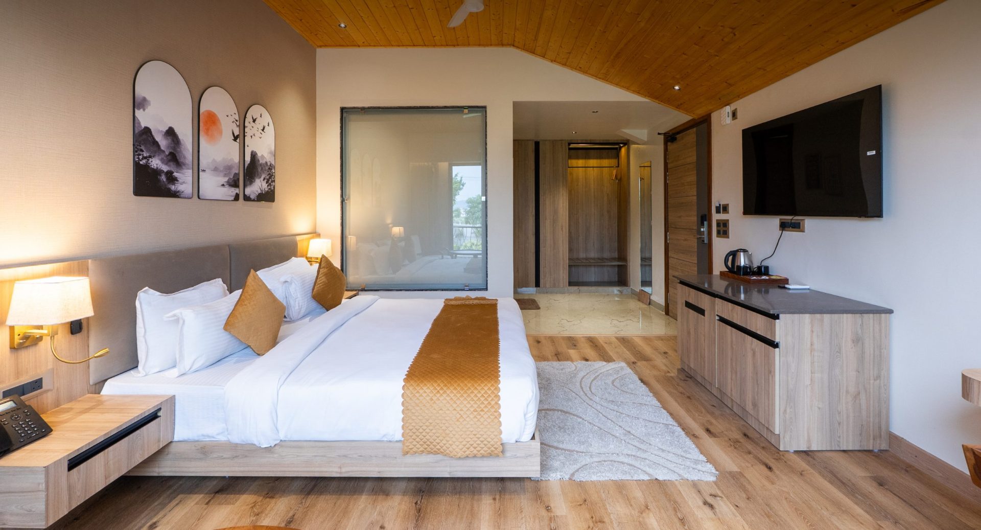 Hotel room with a large bed, tan headboard, and amber throw pillows; wooden floors and ceiling create a warm vibe, with a flat-screen TV on the right.