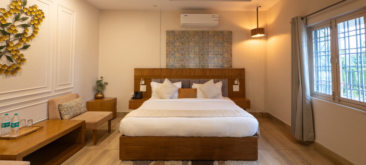 Dehradun – Deluxe Room 5 Hotel room with a large wooden-bed, white linens, and a patterned headboard, plus a seating area to the left.