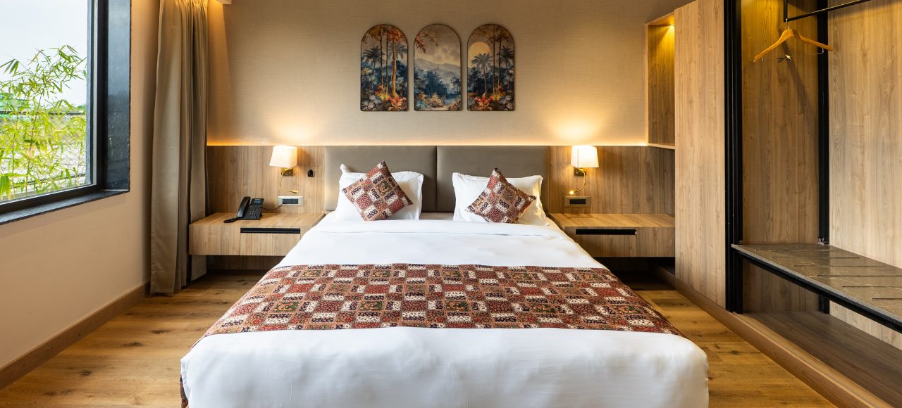 Hotel bedroom with a king bed, white linens, patterned runner and throw pillows, and two nightstands with lamps on either side of the headboard.
