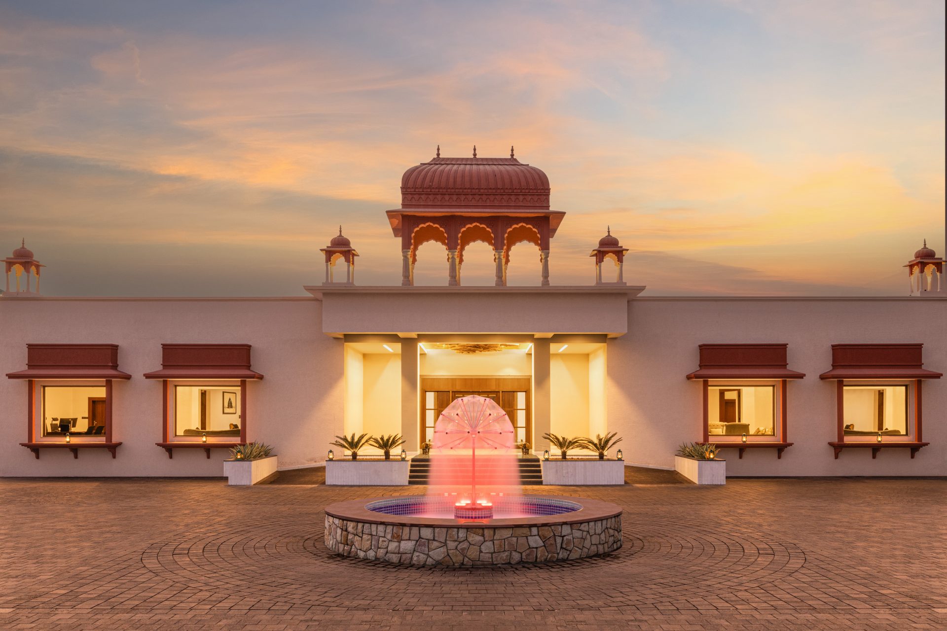 Symmetrical building with an ornate domed pavilion and arched colonnade, pink-lit fountain in the foreground at sunset