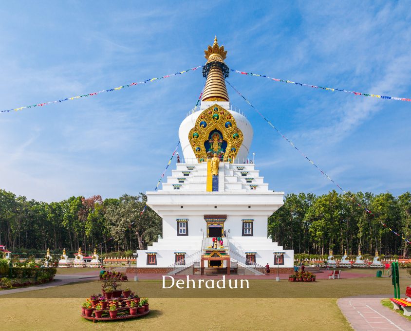 White multi-tiered Buddhist stupa with gold accents in a landscaped park, prayer flags strung overhead against a blue sky, location Dehradun implied by text at bottom.