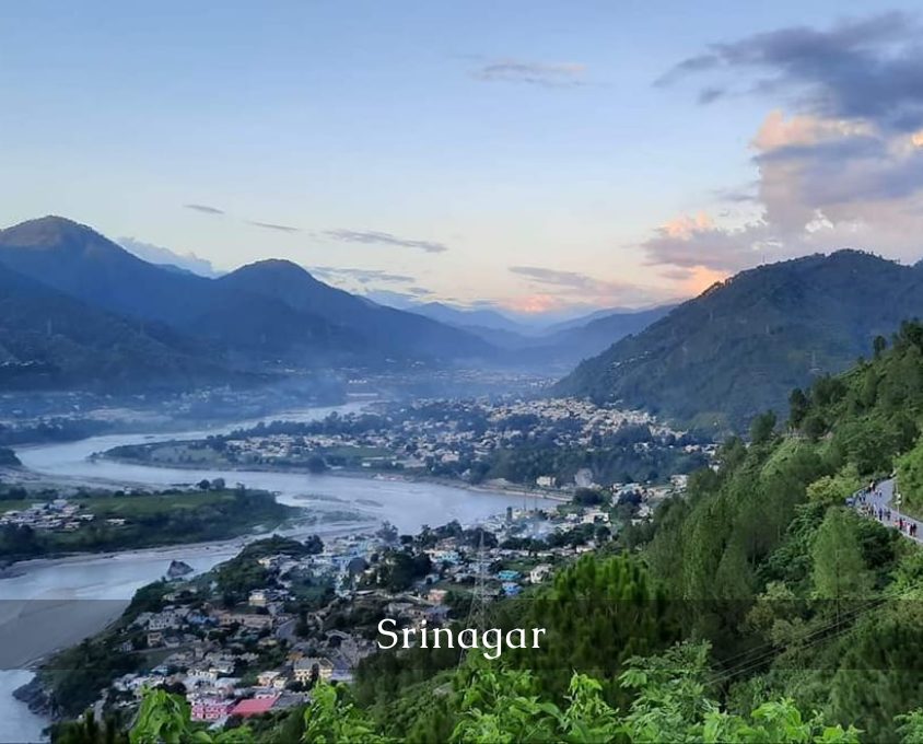 Panoramic view of Srinagar with a winding river through the town and surrounding green hills at sunset/dusk.