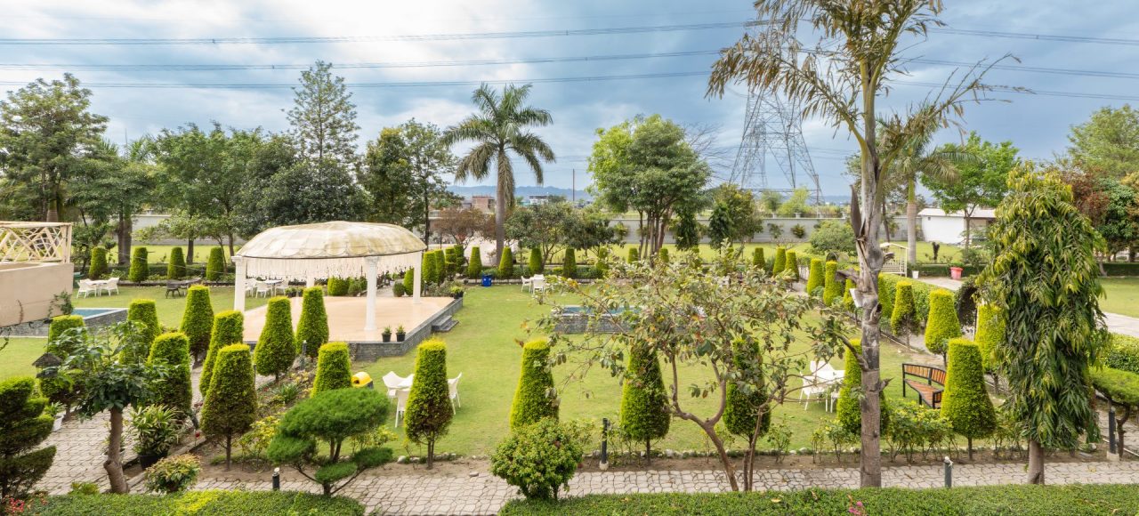 A manicured garden courtyard with a domed white gazebo, trimmed topiary, white chairs, and a paved walkway; power lines and a tall transmission tower are visible in the distance under a cloudy sky.