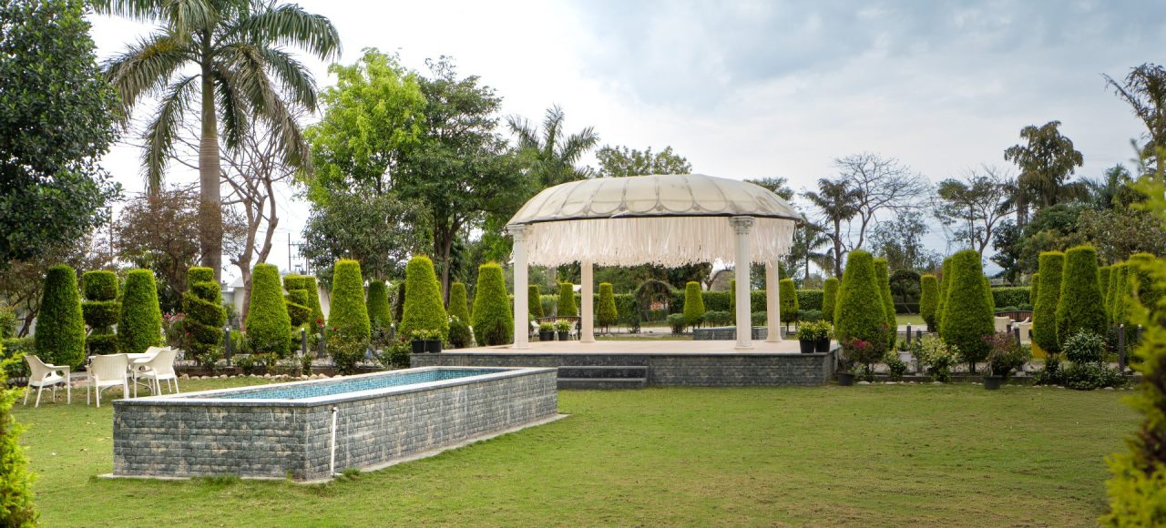 A manicured garden with a white gazebo, trimmed topiary, and a surrounding stone pool, under a cloudy sky.