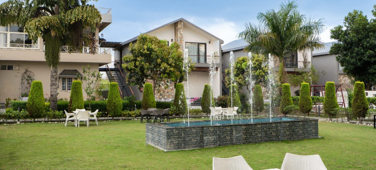 Courtyard of a resort with a stone-edged rectangular fountain spraying jets, surrounded by white tables and chairs, trimmed hedges, and palm trees in the background.