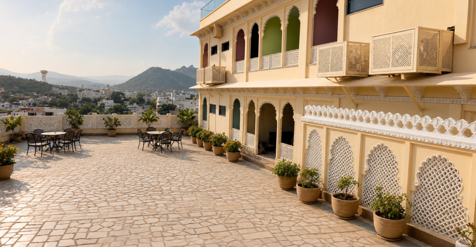Terrace with tan tables and chairs, potted plants, and a beige building with ornate arches, overlooking a town and hills.