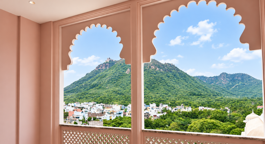 jüSTa Luxé – Monsoon Palace, Udaipur Pink-walled room with scalloped-arch window frames overlooking a green hillside town under a blue sky.