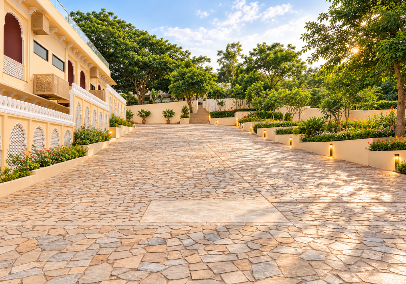 Wide stone-paved courtyard stretching toward steps, with a yellow decorative building on the left and lush trees on the right.
