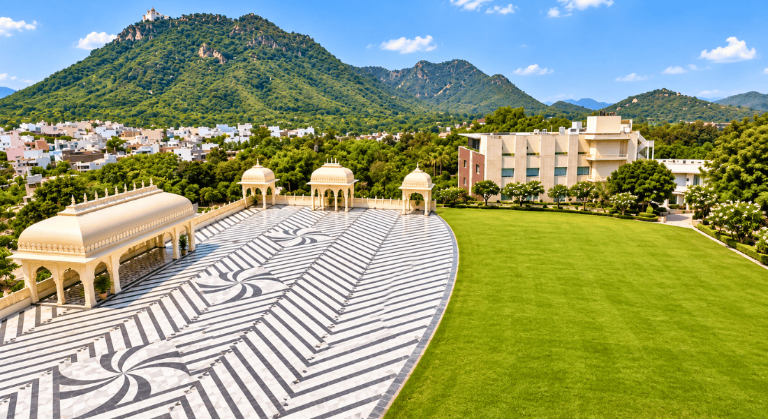 Monsoon 6 A decorative marble plaza with black-and-white geometric tiling, covered pavilions, and a large green lawn against a hillside town backdrop.