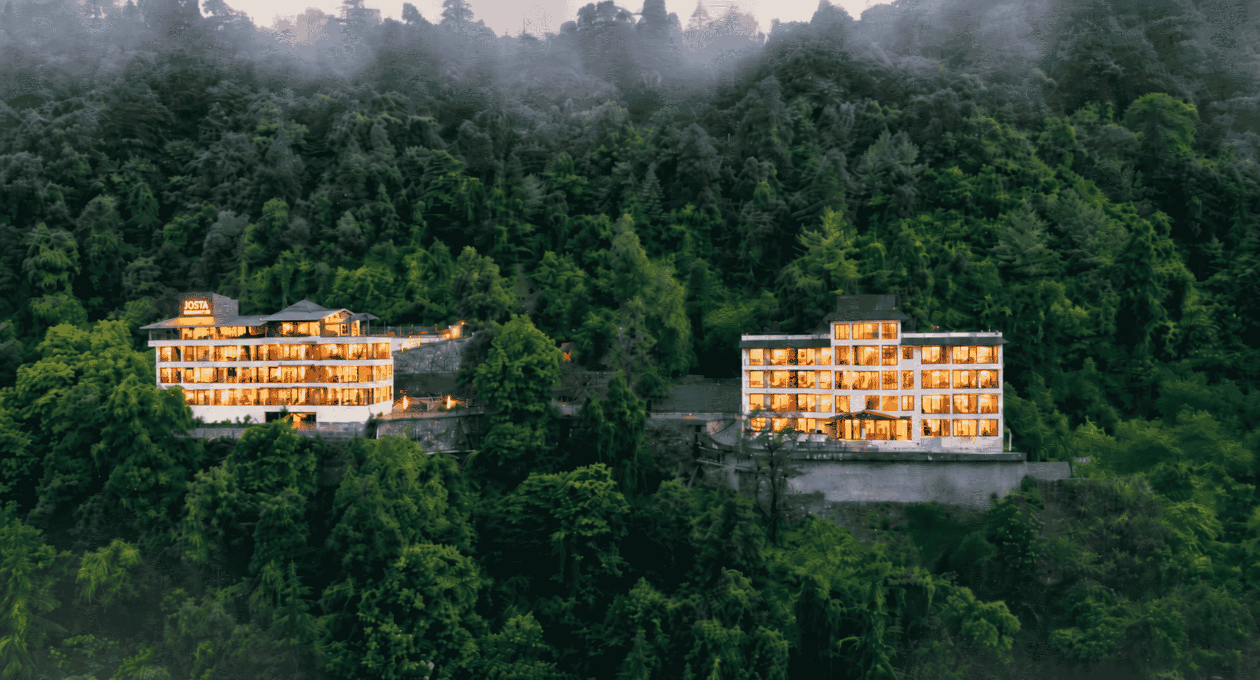 Two modern glass-fronted buildings with warm interior lights nestled in a dense green forest hillside at dusk/or evening.