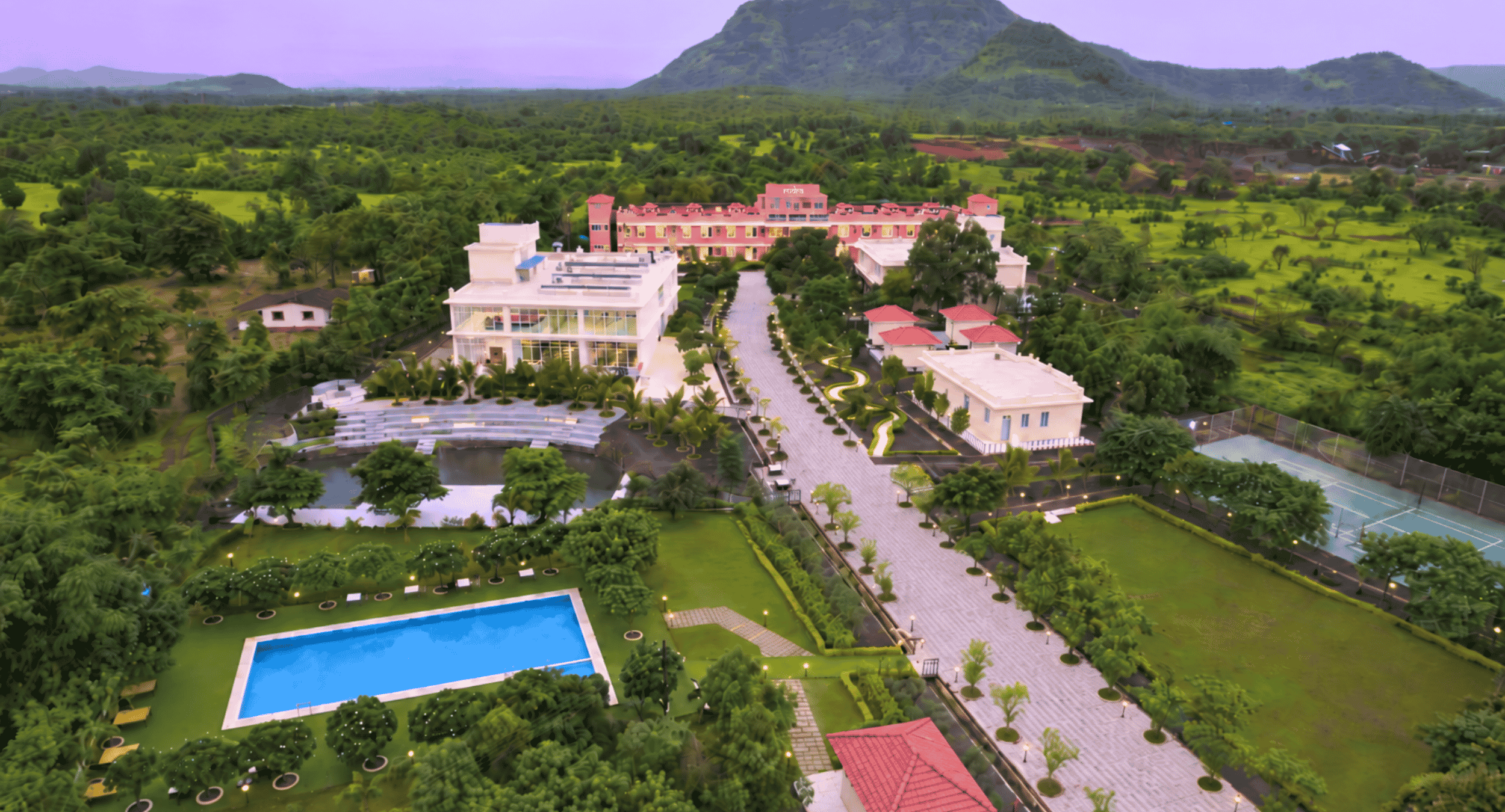 Aerial view of a resort with white modern buildings, a large rectangular swimming pool, palm trees, and manicured gardens set against green fields and a distant mountain range under a purple sky.